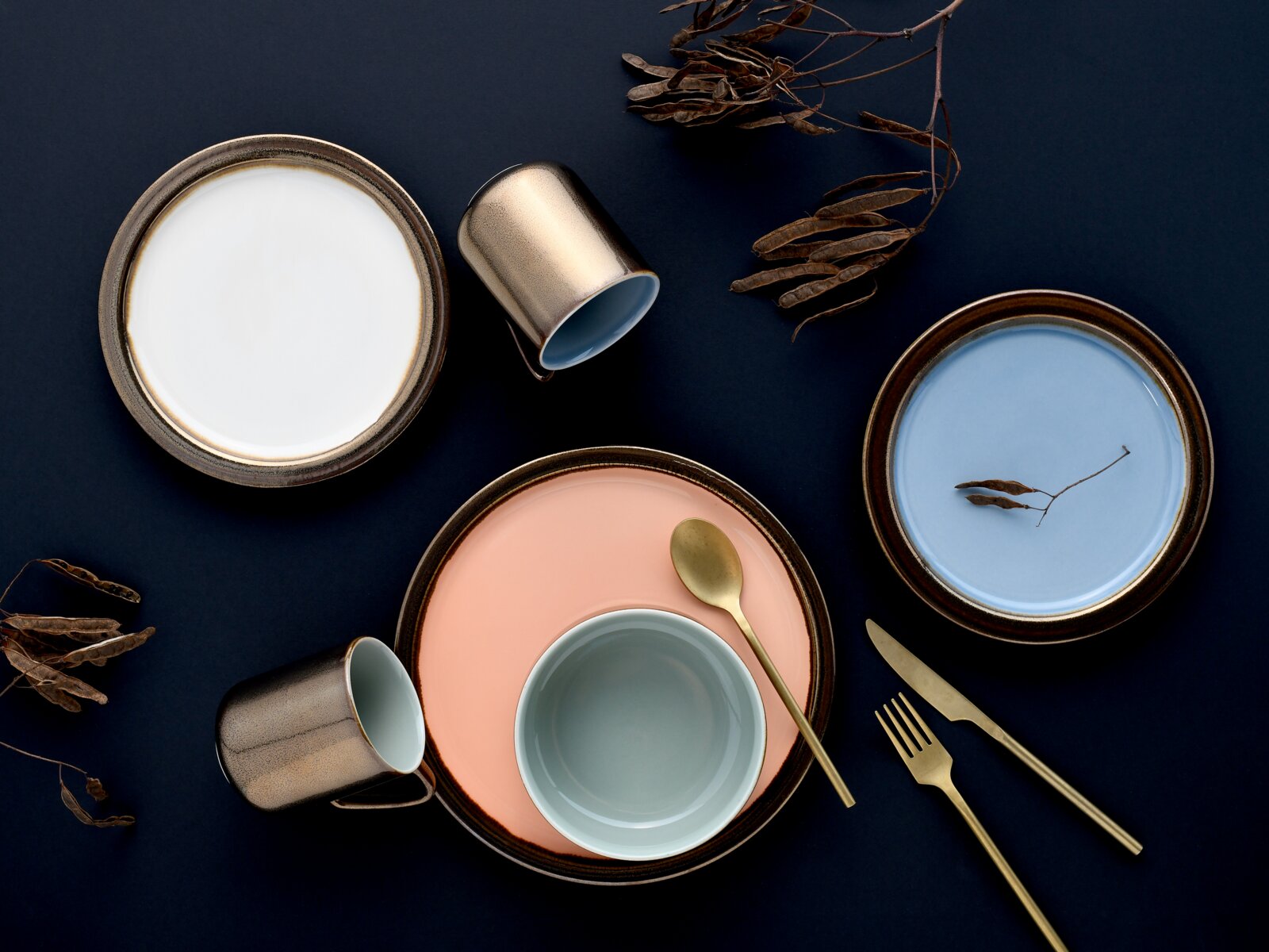 Keramikgeschirr-Set in Pastellfarben mit Tellern, Schale, zwei Tassen und goldenem Besteck auf dunkelblauem Hintergrund, Draufsicht (Flatlay)