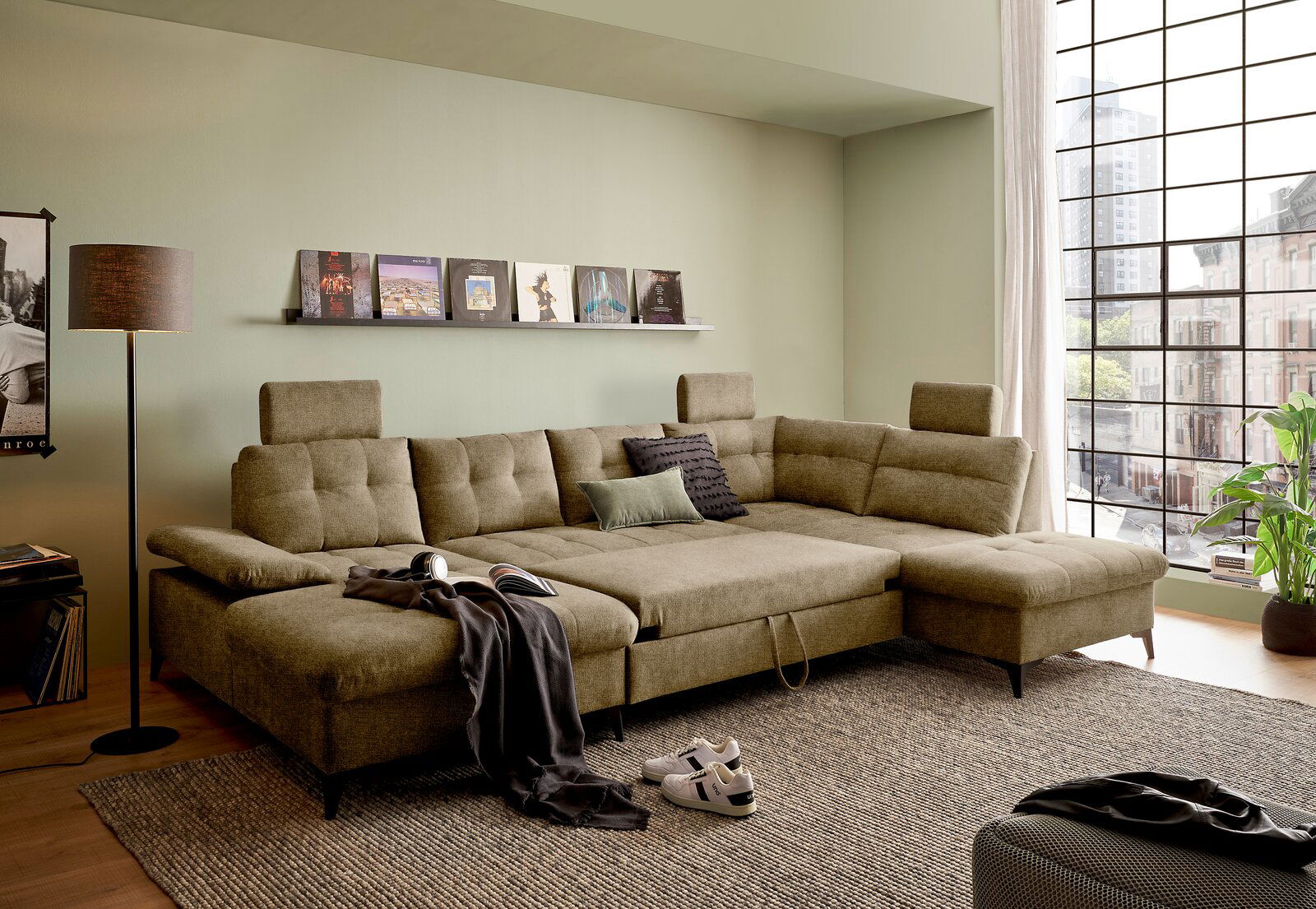 Großes, modernes Ecksofa in einem Wohnzimmer mit grünen Wänden, dekoriert mit Kissen und Decken, aus der Perspektive von vorne links betrachtet.