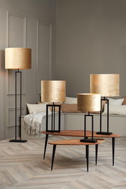 Goldene zylindrische Lampenschirme in verschiedenen Größen auf schwarzen Metallständern, fotografiert in einem modernen Wohnzimmer mit Holzboden und neutralen Wänden.