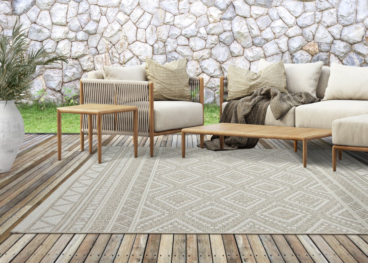 Moderner Outdoor-Teppich auf einer Holzterrasse mit stilvollen Gartenmöbeln, im Hintergrund eine Steinmauer.