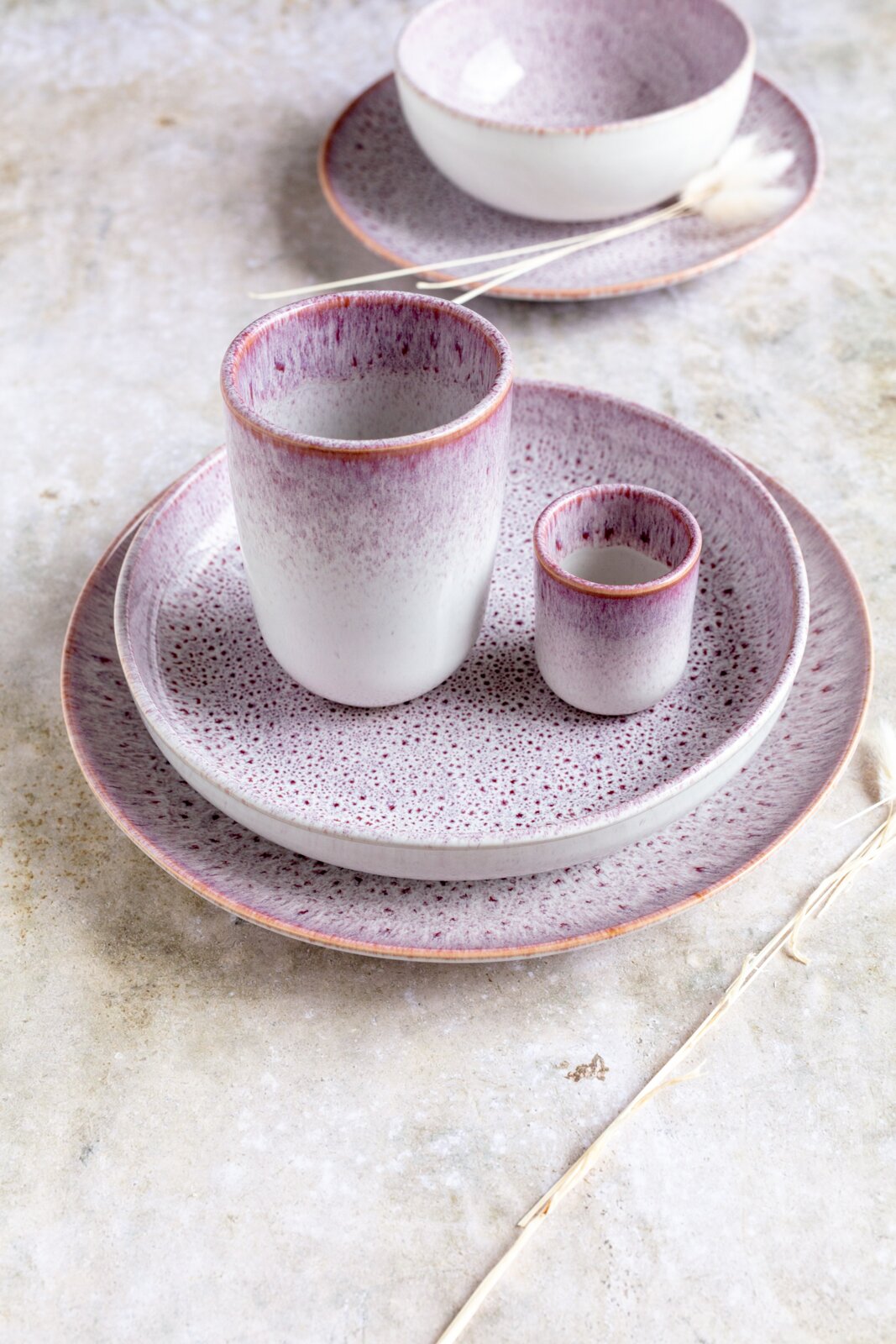 Keramikgeschirr-Set in Weiß mit violett gesprenkelter Glasur: großer und kleiner Becher auf zwei Tellern, im Hintergrund Schale auf Teller; fotografiert in leicht erhöhter, schräger Vorderansicht.