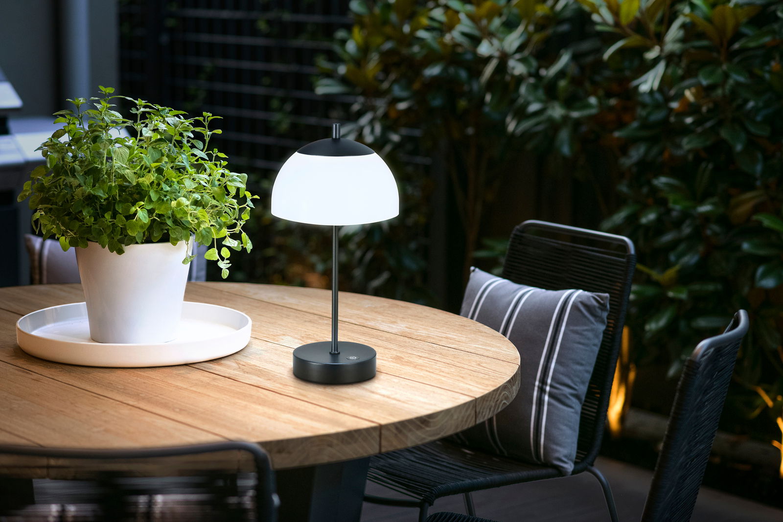 Akku-Tischleuchte RIVA auf einem runden Holztisch im Freien, neben einem weißen Blumentopf mit grüner Pflanze, aus seitlicher Perspektive fotografiert.