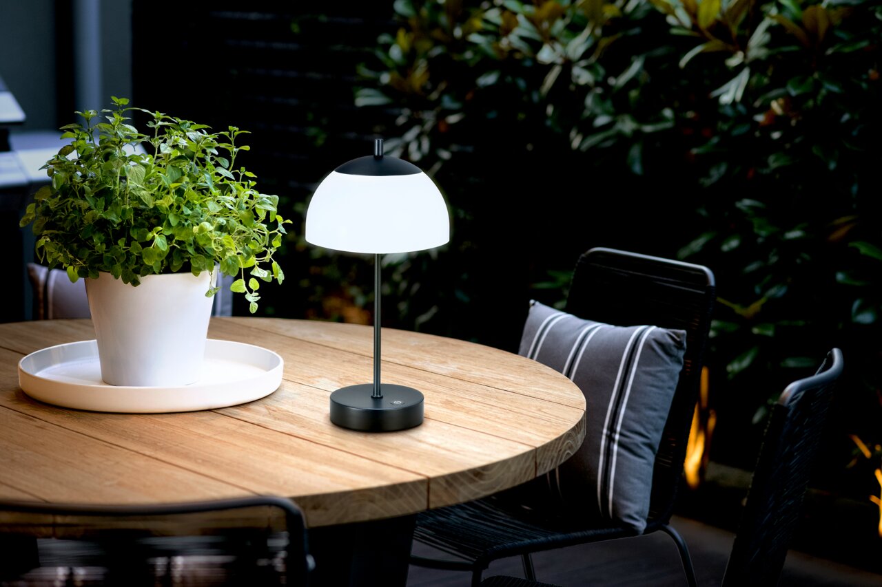 De RIVA-tafellamp op accu op een ronde houten tafel buiten, naast een witte bloempot met een groene plant, gefotografeerd vanuit een zijdelingse hoek.
