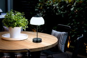 De RIVA-tafellamp op accu op een ronde houten tafel buiten, naast een witte bloempot met een groene plant, gefotografeerd vanuit een zijdelingse hoek.