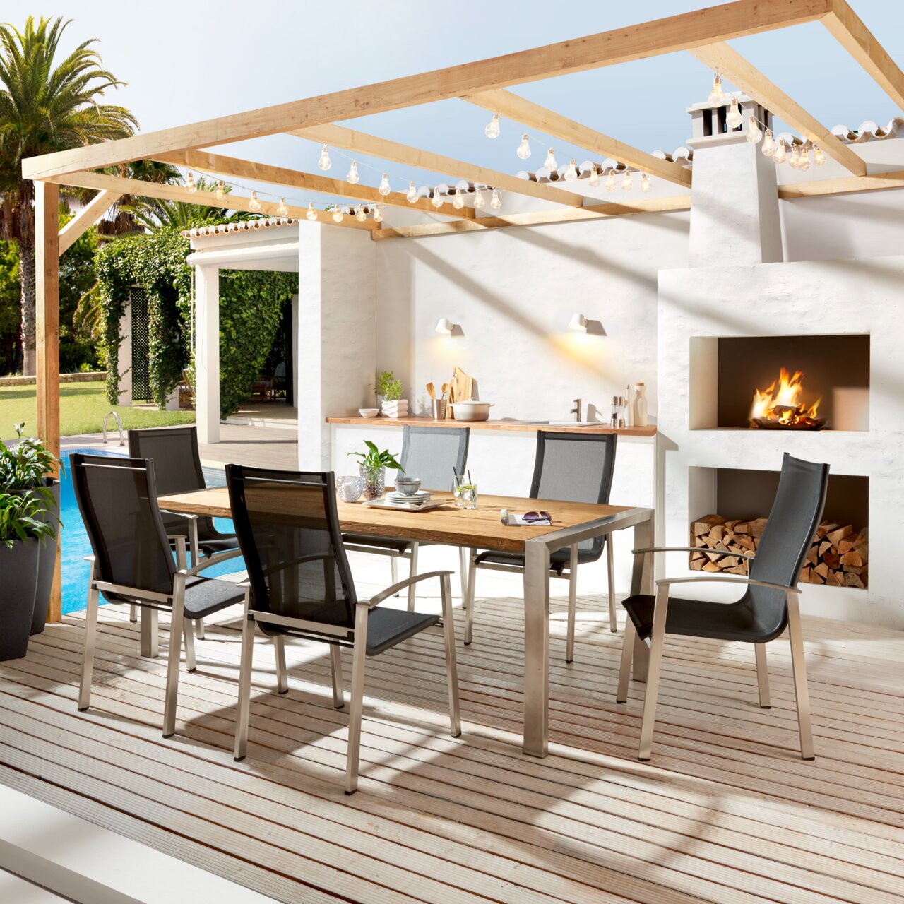 Moderner Gartentisch mit Holzplatte und Metallgestell, umgeben von sechs stilvollen Stühlen auf einer Terrasse mit Blick auf einen Pool und eine Außenküche.