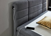 Graues Boxspringbett mit gepolstertem Kopfteil, USB-Ladeanschluss und goldfarbenem Kissen, seitliche Perspektive