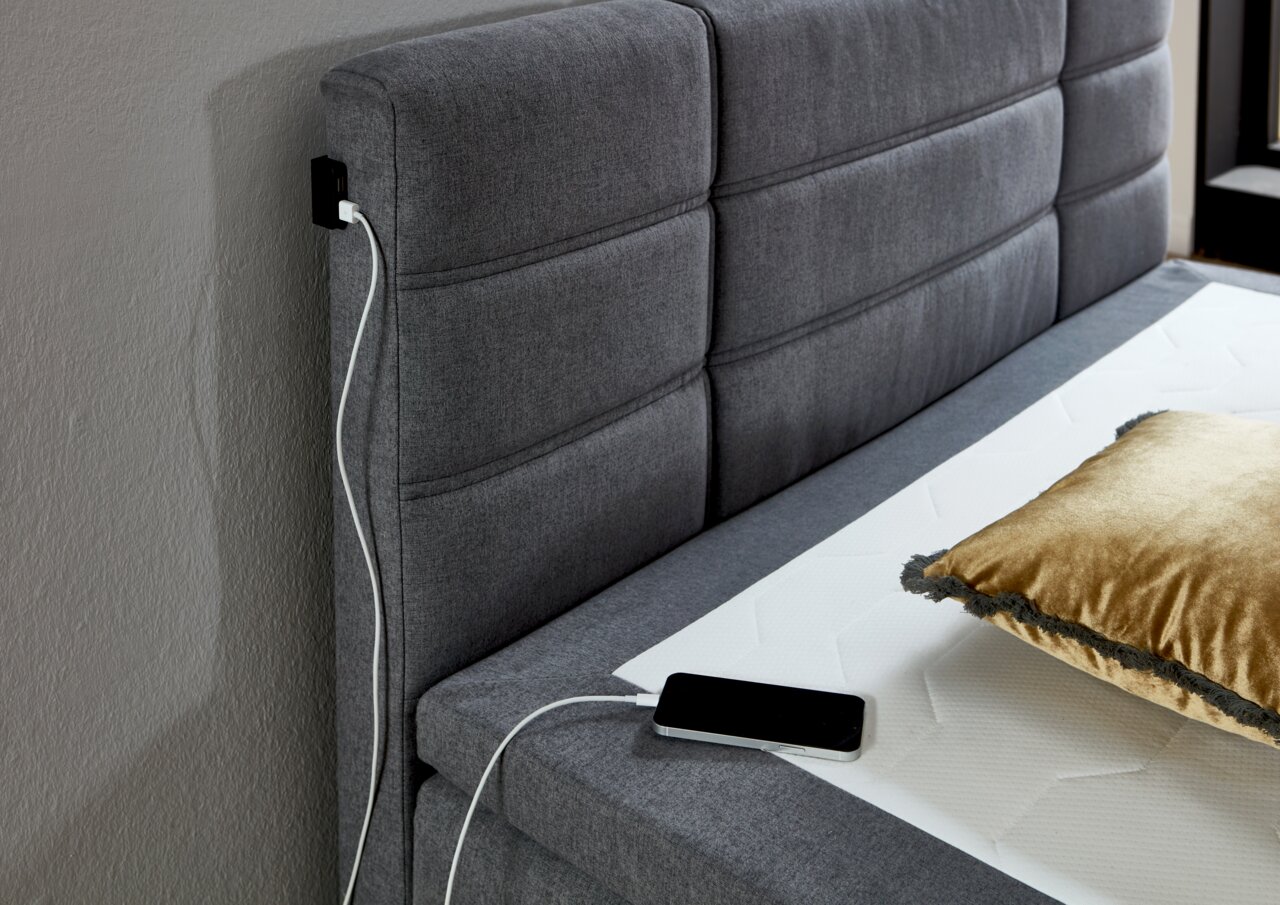 Graues Boxspringbett mit gepolstertem Kopfteil, USB-Ladeanschluss und goldfarbenem Kissen, seitliche Perspektive
