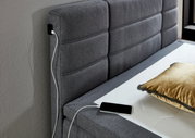 Graues Boxspringbett mit gepolstertem Kopfteil, USB-Ladeanschluss und goldfarbenem Kissen, seitliche Perspektive