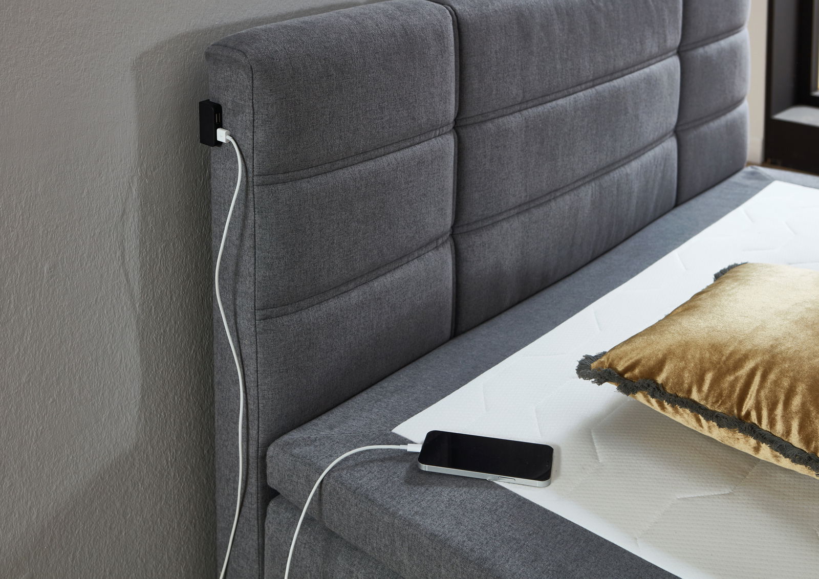 Graues Boxspringbett mit gepolstertem Kopfteil, USB-Ladeanschluss und goldfarbenem Kissen, seitliche Perspektive
