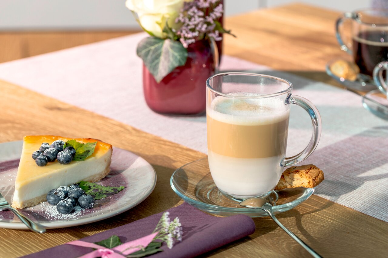 Leonardo Untertasse NAPOLI Untertasse Napoli 14,3cm mit Glas, gefüllt mit Latte Macchiato, von oben fotografiert, neben einem Stück Käsekuchen mit Blaubeeren und einem Keks.