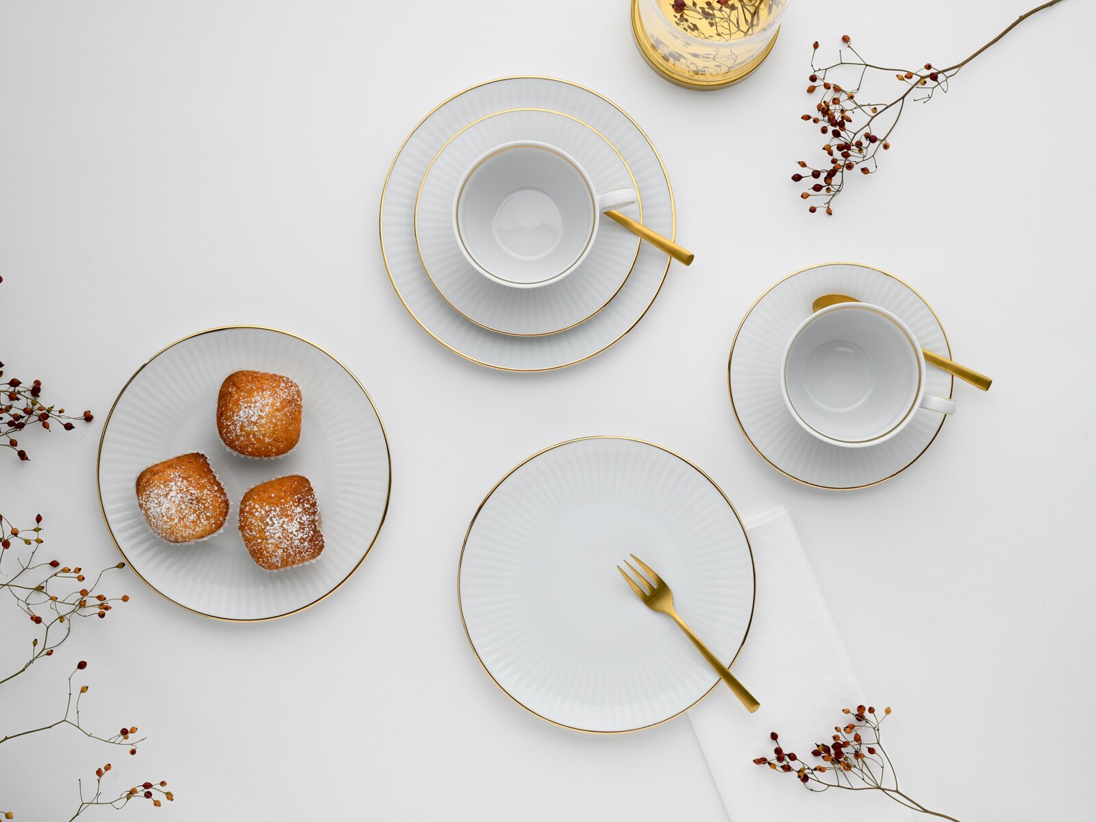 Draufsicht auf elegantes weißes Porzellan-Kaffeegeschirr mit goldenen Rändern: zwei Tassen auf Untertassen, mehrere Teller, goldfarbene Besteckteile und drei kleine Kuchen auf einem Teller auf hellem Hintergrund.