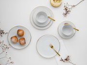 Draufsicht auf elegantes weißes Porzellan-Kaffeegeschirr mit goldenen Rändern: zwei Tassen auf Untertassen, mehrere Teller, goldfarbene Besteckteile und drei kleine Kuchen auf einem Teller auf hellem Hintergrund.