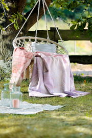Wohndecke in sanften Pastelltönen auf einer Hängeschaukel im Garten, umgeben von grüner Natur und dekorativen Lichtern, aus der Perspektive von vorne.