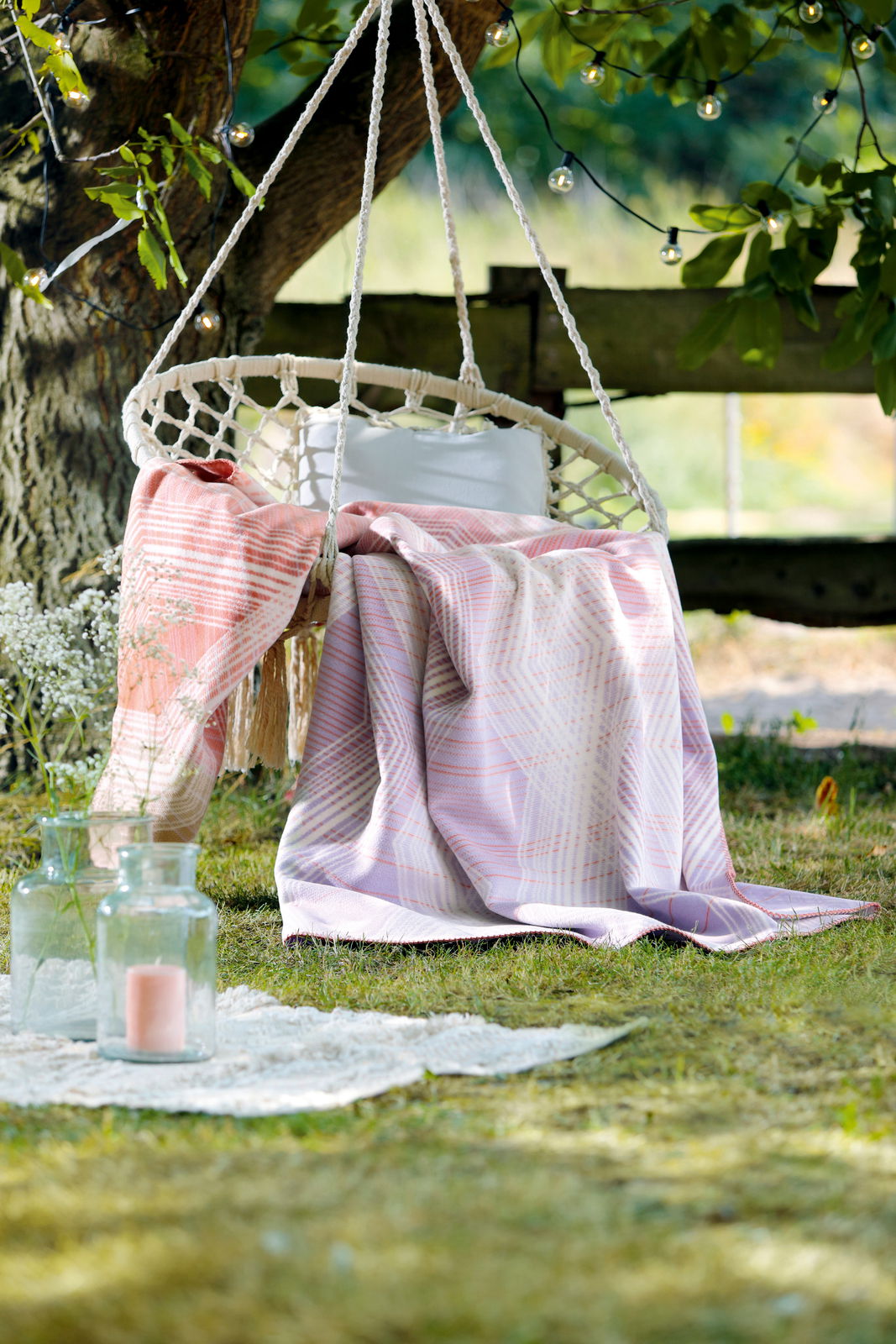 Wohndecke in sanften Pastelltönen auf einer Hängeschaukel im Garten, umgeben von grüner Natur und dekorativen Lichtern, aus der Perspektive von vorne.