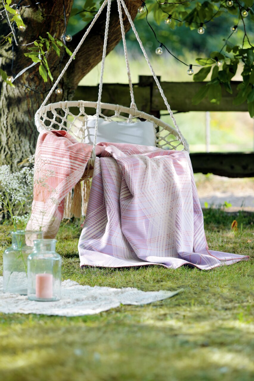biederlack Woonkleed TREND Een deken in zachte pasteltinten op een hangschommel in de tuin, omringd door groene natuur en sfeerverlichting, gezien van voren.