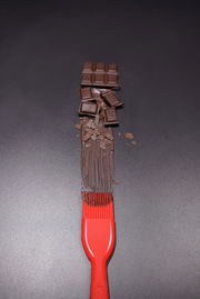 Roter Silikon-Backpinsel von oben, der geschmolzene Schokolade auf einer dunklen Oberfläche verteilt, mit Schokoladenstücken im Hintergrund.