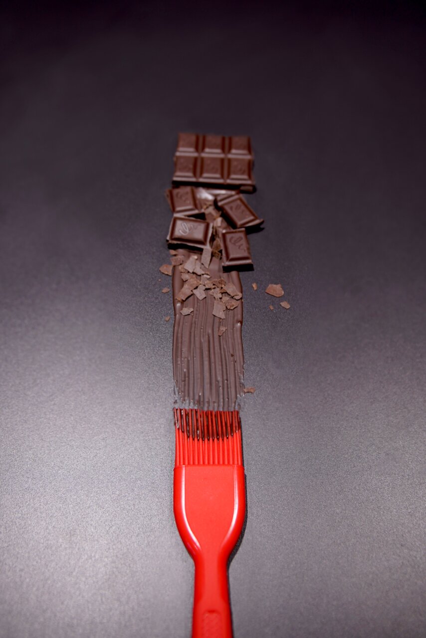 Dr. Oetker Silikon-Backpinsel Roter Silikon-Backpinsel von oben, der geschmolzene Schokolade auf einer dunklen Oberfläche verteilt, mit Schokoladenstücken im Hintergrund.