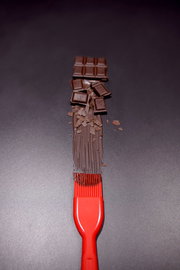 Roter Silikon-Backpinsel von oben, der geschmolzene Schokolade auf einer dunklen Oberfläche verteilt, mit Schokoladenstücken im Hintergrund.