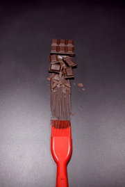 Roter Silikon-Backpinsel von oben, der geschmolzene Schokolade auf einer dunklen Oberfläche verteilt, mit Schokoladenstücken im Hintergrund.