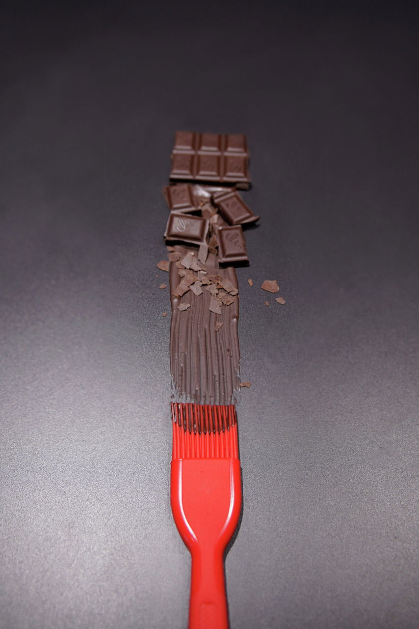 Roter Silikon-Backpinsel von oben, der geschmolzene Schokolade auf einer dunklen Oberfläche verteilt, mit Schokoladenstücken im Hintergrund.