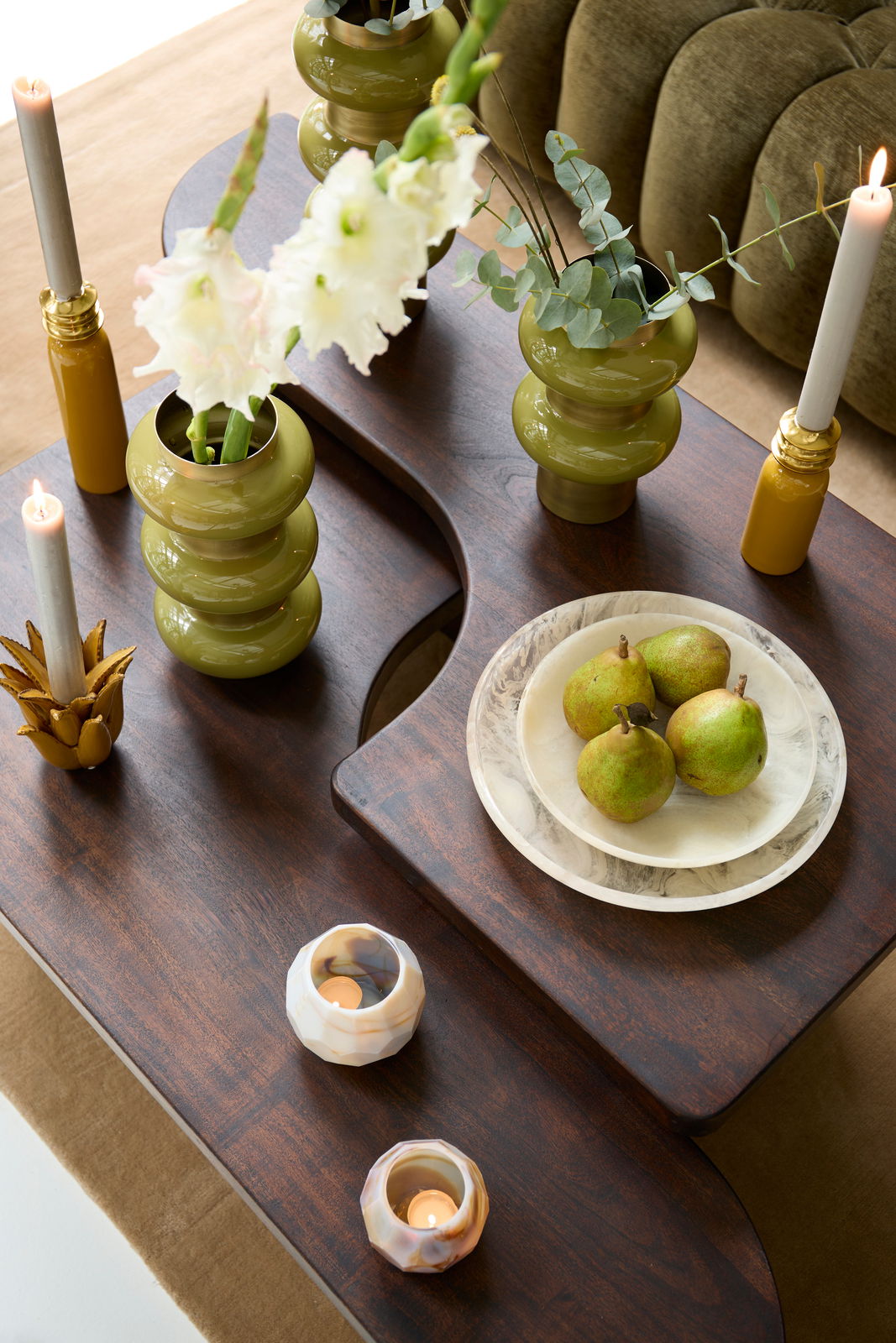 Vogelperspektive auf einen stilvollen Couchtisch mit dunkler Holzoberfläche, dekoriert mit grünen Vasen, Kerzen und einem Teller mit Birnen.