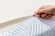 Nahaufnahme eines Matratzentoppers mit Reißverschluss, der von einer Hand geöffnet wird. Der Topper hat ein gestreiftes Muster und zeigt die weiche, gepolsterte Oberfläche.