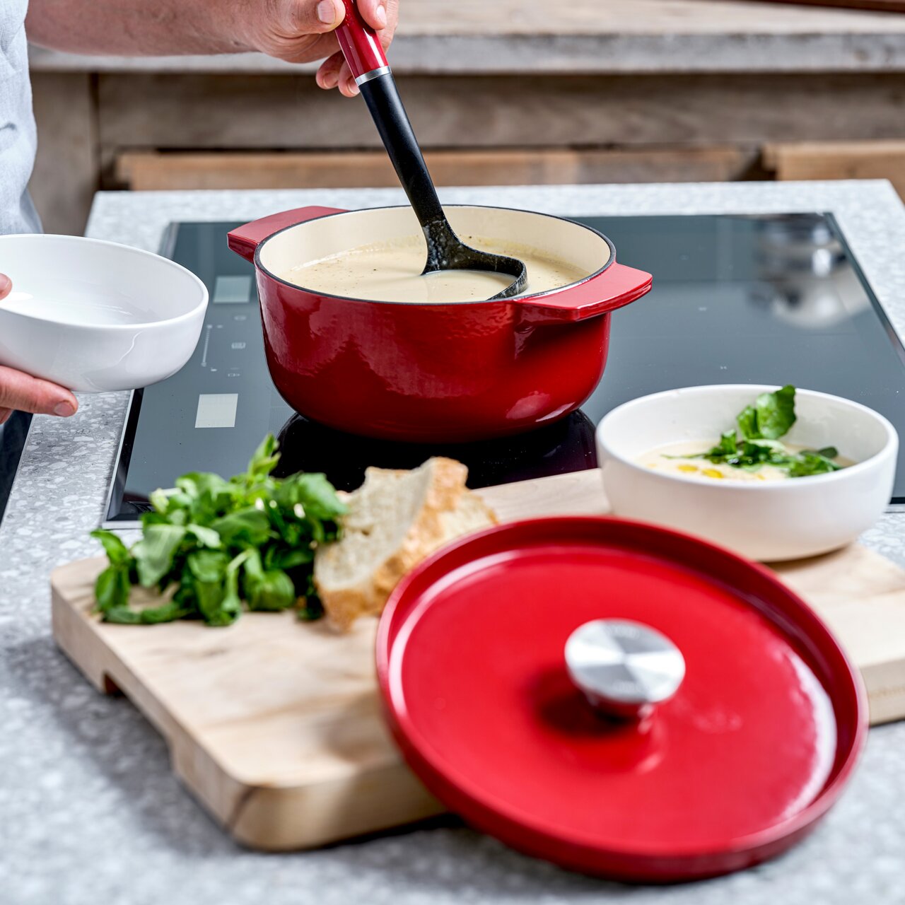 KitchenAid Bräter Roter runder Gusseisen Bräter von KitchenAid auf einem Herd, gefüllt mit Suppe. Im Vordergrund liegt der Deckel auf einem Schneidebrett mit Brot und Kräutern. Perspektive von schräg oben.