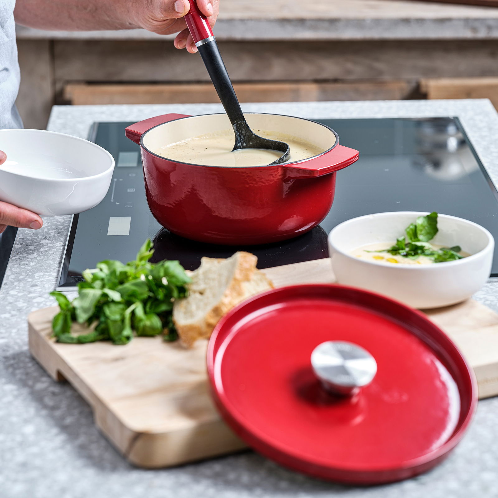 Roter runder Gusseisen Bräter von KitchenAid auf einem Herd, gefüllt mit Suppe. Im Vordergrund liegt der Deckel auf einem Schneidebrett mit Brot und Kräutern. Perspektive von schräg oben.