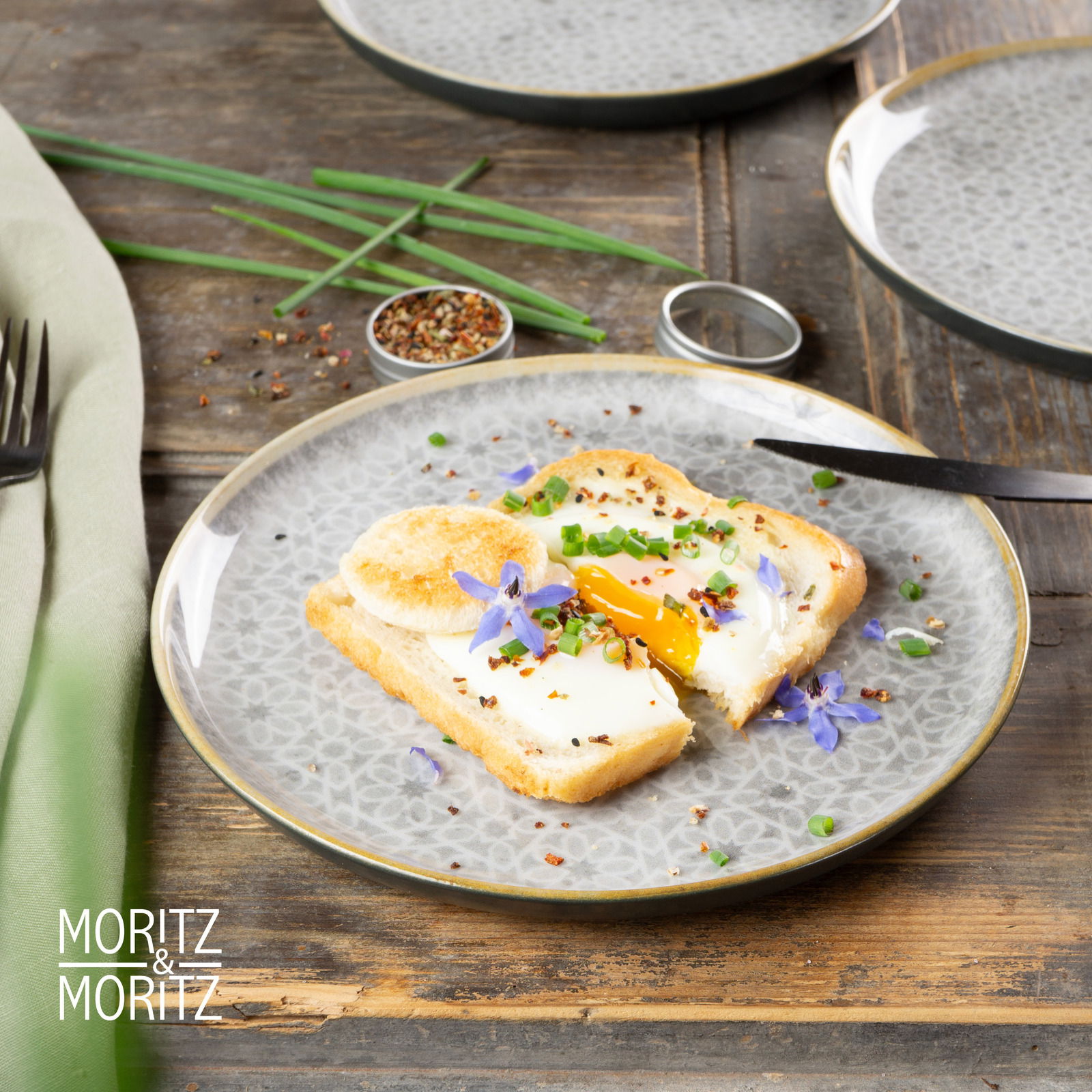 Graues Geschirrset Terra von Moritz & Moritz, 18-teilig, mit einem Teller im Vordergrund, darauf ein Toast mit Spiegelei und essbaren Blüten, aus der Vogelperspektive fotografiert.