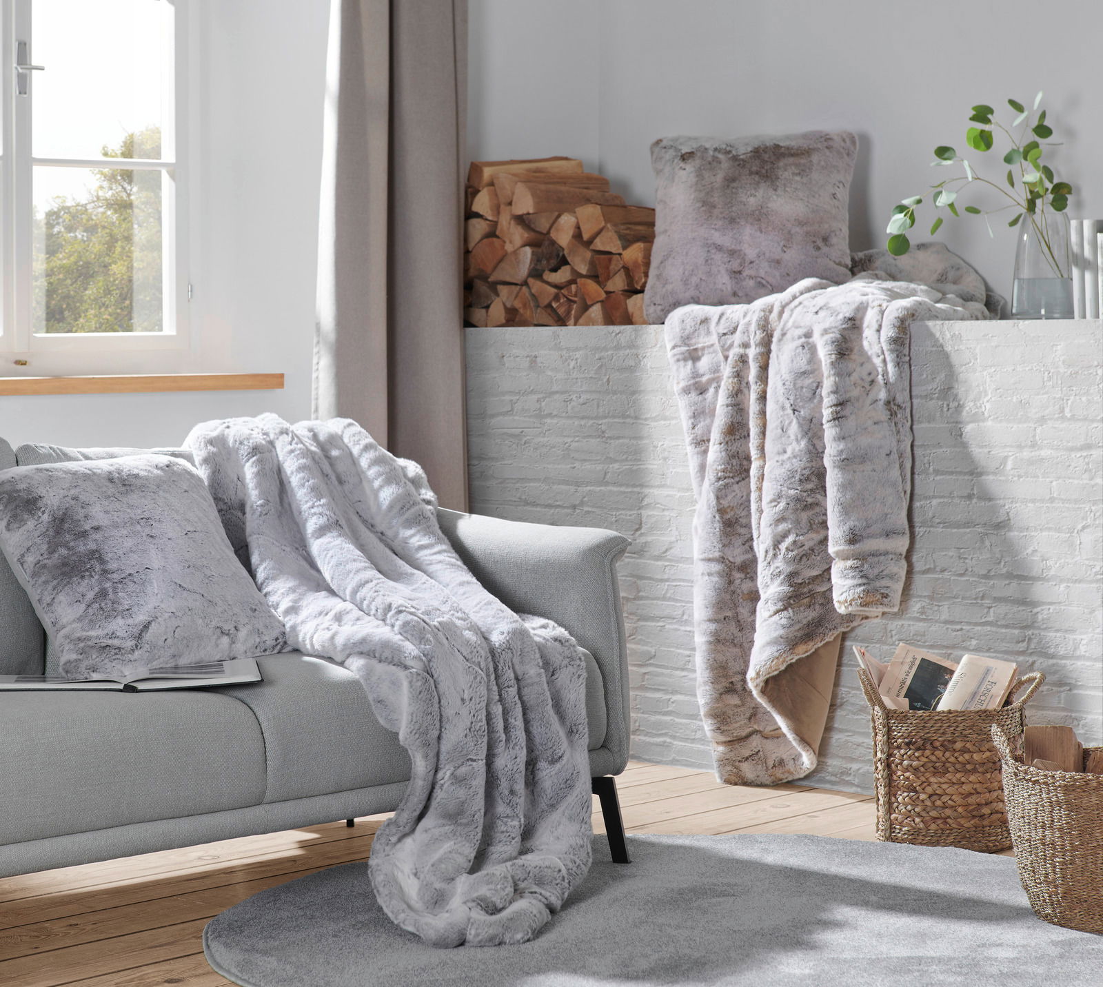 Gemütliche Wohnzimmer-Szene mit einer silberfuchsfarbenen Felldecke und passenden Kissen auf einem grauen Sofa, im Hintergrund ein Kamin mit Holzscheiten und Pflanzen, seitliche Perspektive.