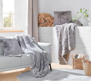 Gemütliche Wohnzimmer-Szene mit einer silberfuchsfarbenen Felldecke und passenden Kissen auf einem grauen Sofa, im Hintergrund ein Kamin mit Holzscheiten und Pflanzen, seitliche Perspektive.