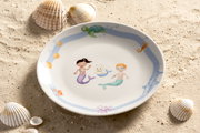 Kinderteller mit Meerjungfrauenmotiv, umgeben von Muscheln auf Sand, aus der Vogelperspektive fotografiert.