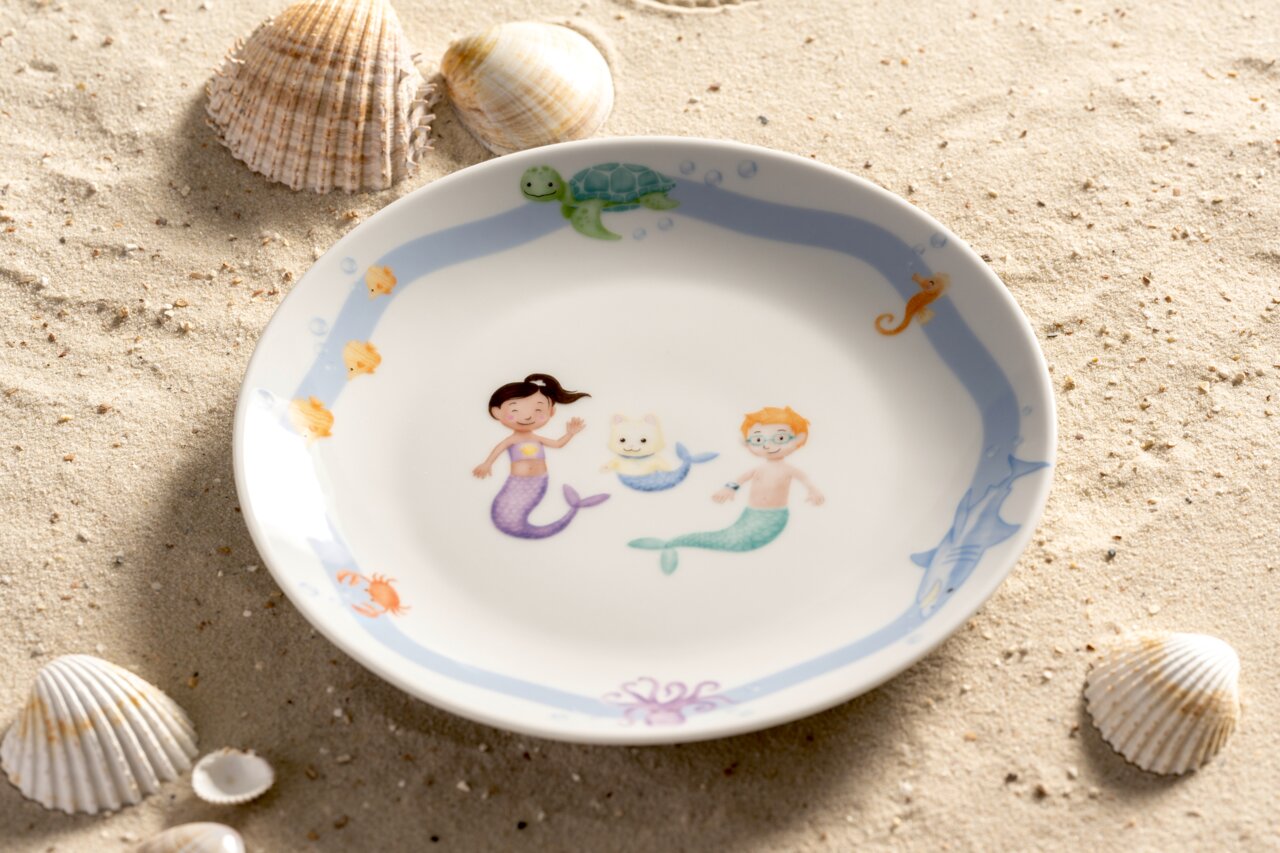 Kinderteller mit Meerjungfrauenmotiv, umgeben von Muscheln auf Sand, aus der Vogelperspektive fotografiert.