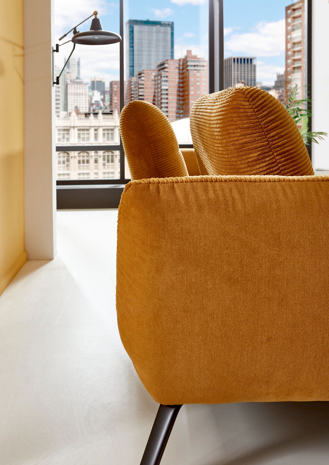 Nahaufnahme einer senfgelben Rundecke mit modernen schwarzen Beinen, im Hintergrund eine städtische Skyline durch ein großes Fenster sichtbar.