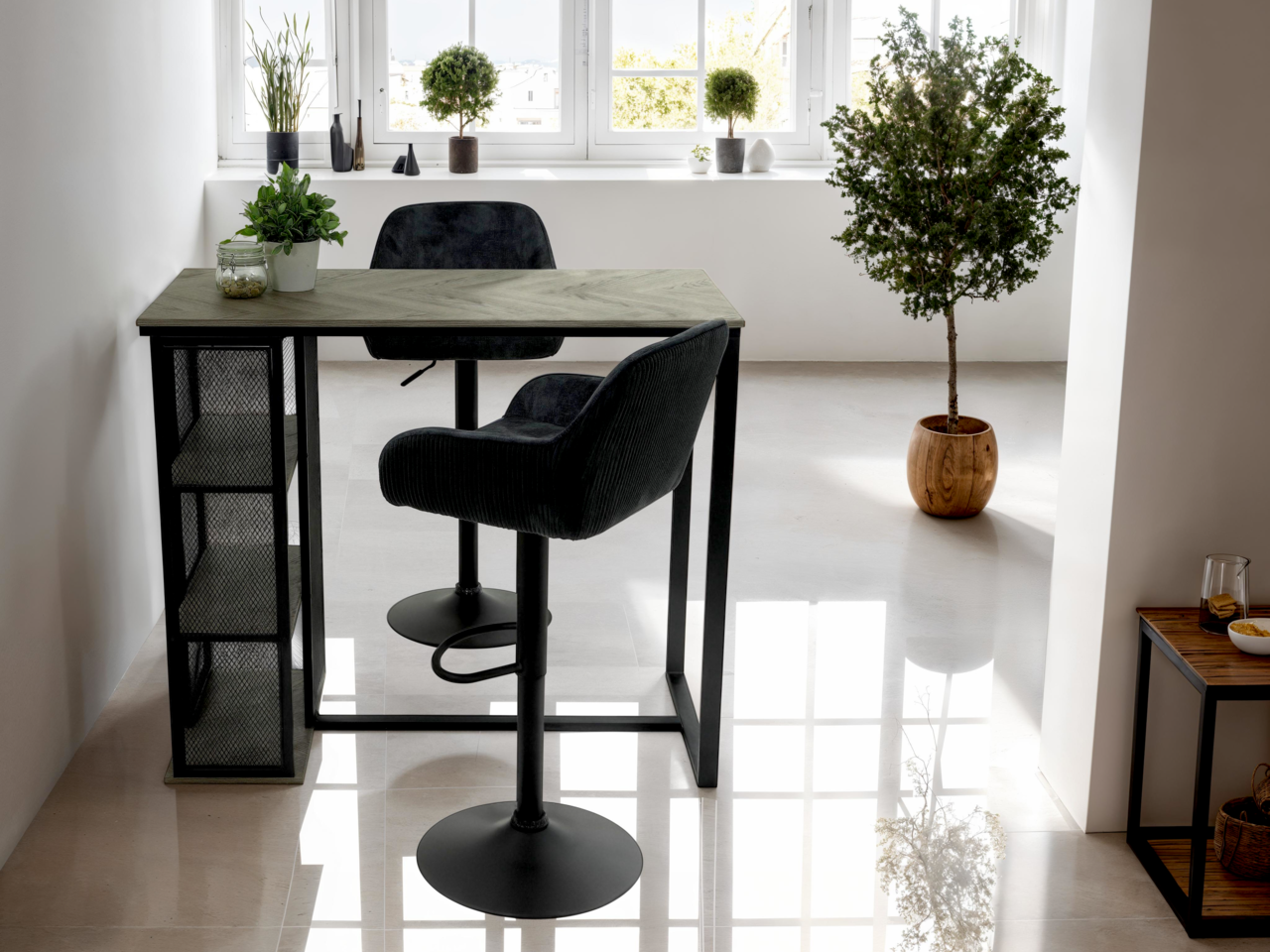 Moderner Bartisch mit schwarzem Metallgestell und Holzplatte, seitlich mit Regalfächern, in einem hellen Raum mit Pflanzen dekoriert, seitliche Perspektive.