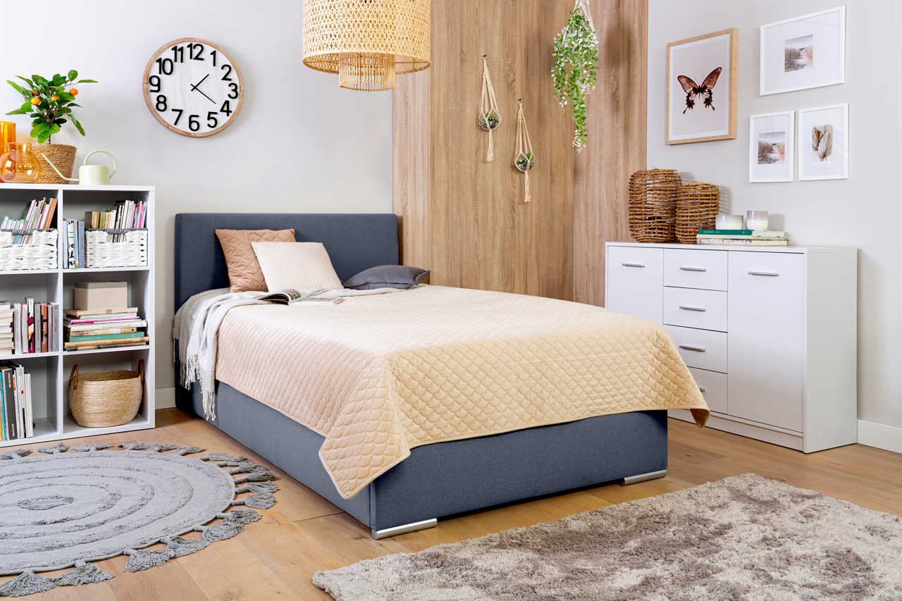 Z2 Boxspringbett TOM Gemütliches Schlafzimmer mit einem Boxbett in Grau, dekoriert mit Kissen und einer beigen Steppdecke, umgeben von modernen Möbeln und Pflanzen, seitliche Perspektive.