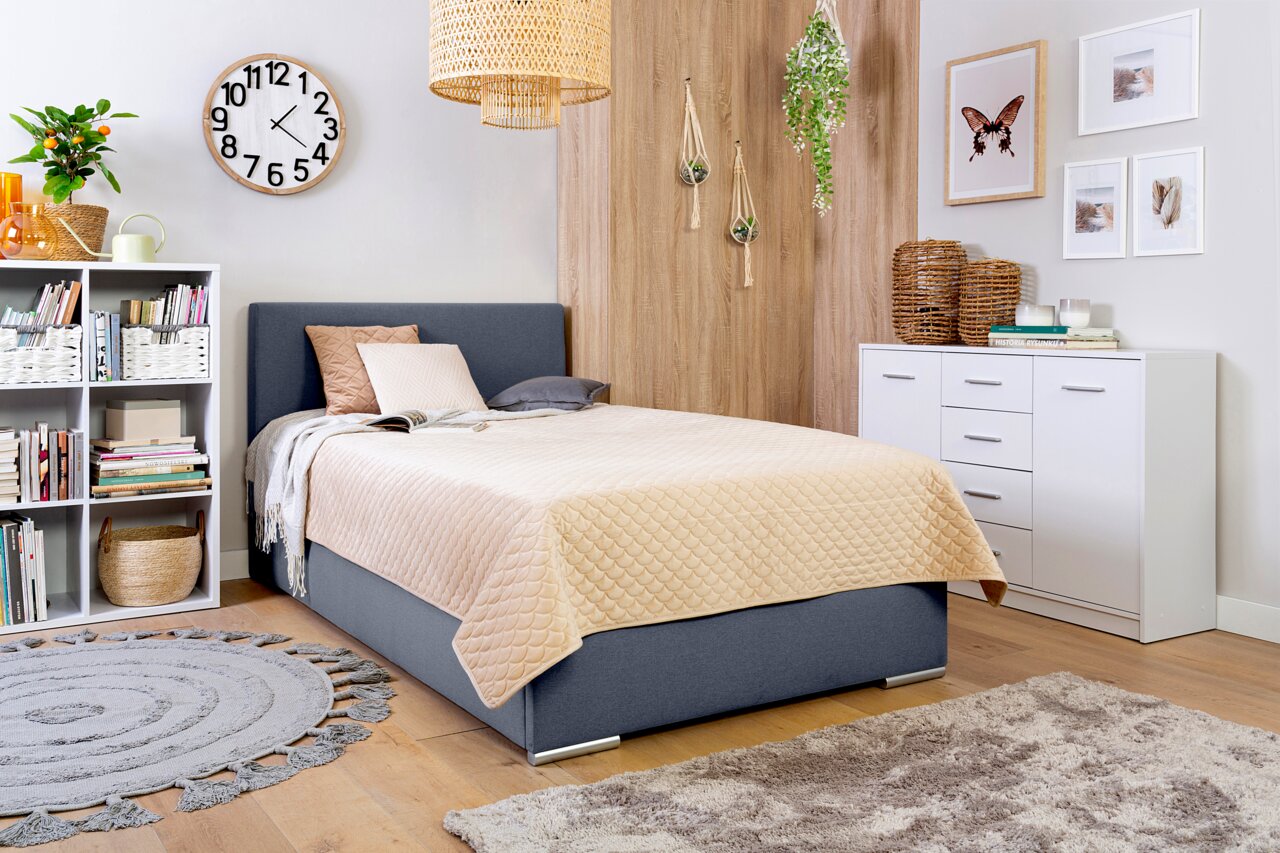 Gezellige slaapkamer met een grijs boxbed, versierd met kussens en een beige dekbed, omringd door moderne meubels en planten, zijaanzicht.