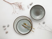 Draufsicht auf ein minimalistisches Keramik-Geschirrset in Grau: zwei Teller mit goldfarbenem Messer und Gabel sowie eine passende Schale auf weißem Hintergrund, dekoriert mit kleinen Steinen und Trockenblumen.