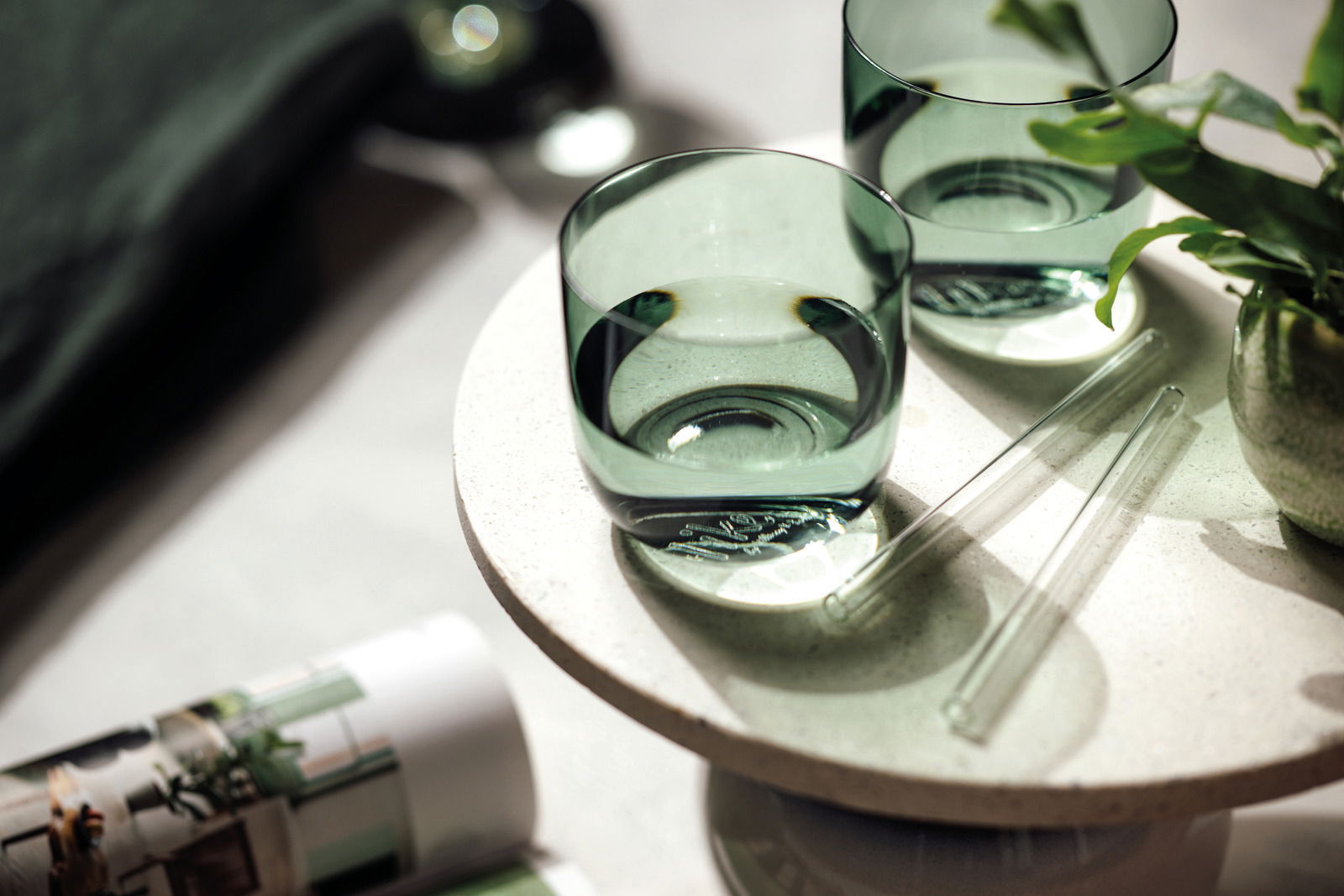 Zwei grüne Wassergläser auf einem runden, hellen Tisch aus der Vogelperspektive, umgeben von Pflanzen und Glasstäben.