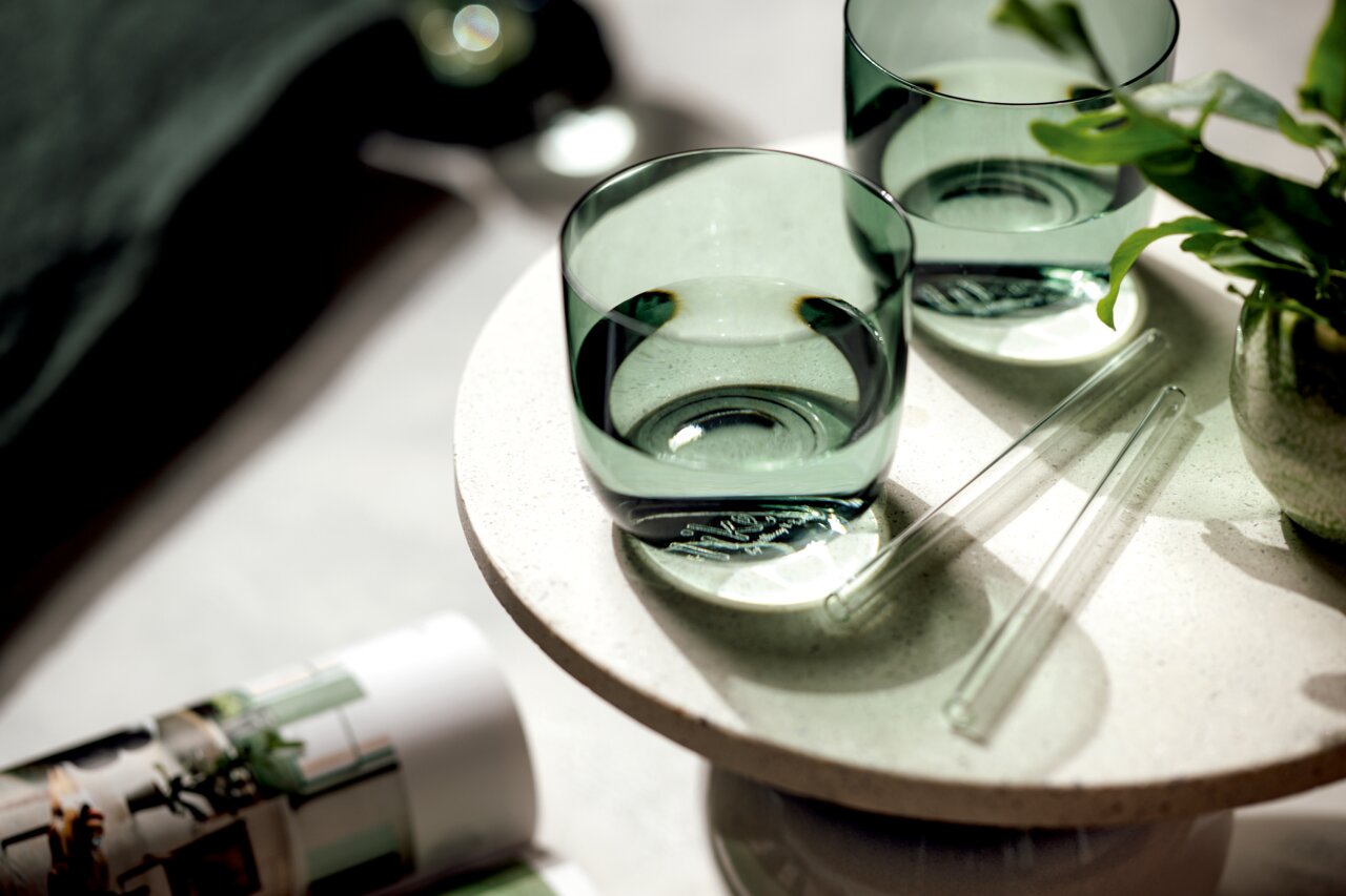 Zwei grüne Wassergläser auf einem runden, hellen Tisch aus der Vogelperspektive, umgeben von Pflanzen und Glasstäben.
