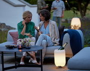 Zwei Frauen sitzen auf einem Sofa im Freien, umgeben von stimmungsvoller Beleuchtung und Dekoration. Im Vordergrund steht ein leuchtender, silberner Atmos-Kühler mit einer Flasche Champagner. Perspektive: seitlich von vorne.