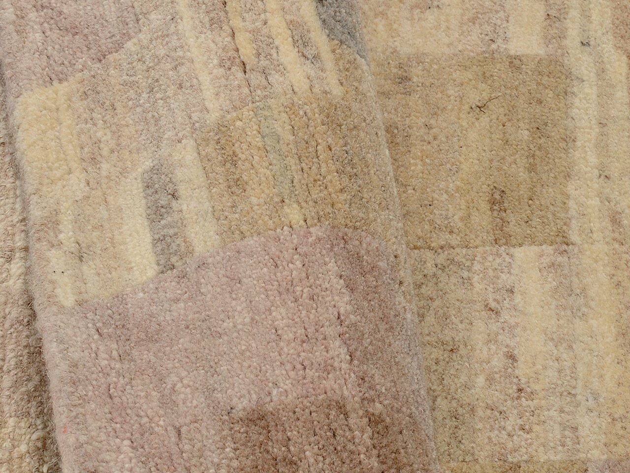 Nahaufnahme eines Wollteppichs mit geometrischem Muster in sanften Beige- und Rosatönen, aus seitlicher Perspektive aufgenommen.