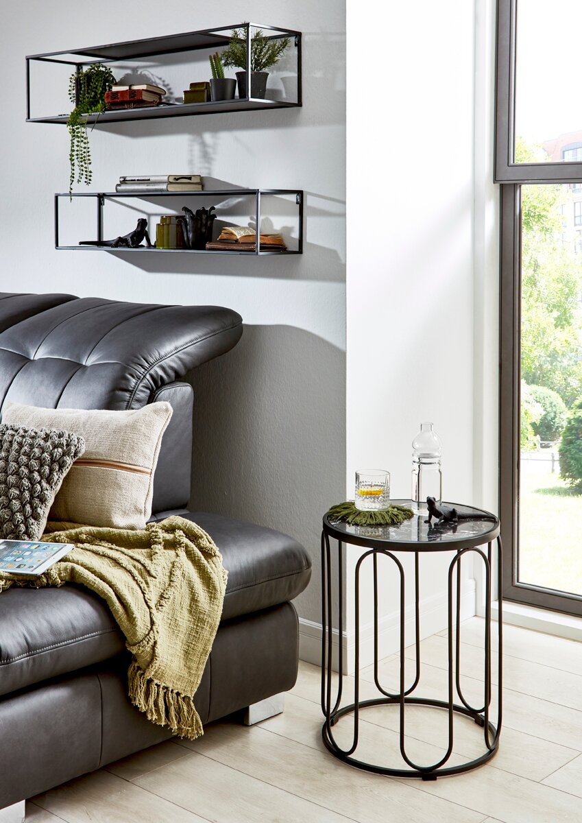 Modernes Wohnzimmer-Setting: runder Beistelltisch mit schwarzem Metallgestell und Glasplatte neben grauem Ledersofa am Fenster, Wandregale im Hintergrund; Perspektive leicht seitlich in Augenhöhe