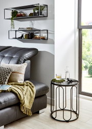 Modernes Wohnzimmer-Setting: runder Beistelltisch mit schwarzem Metallgestell und Glasplatte neben grauem Ledersofa am Fenster, Wandregale im Hintergrund; Perspektive leicht seitlich in Augenhöhe