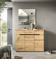 Moderne Holzkommode aus massiver Eiche mit markanter Maserung und schwarzen Griffleisten, frontal in einem hellen Wohnraum unter einem Wandspiegel inszeniert