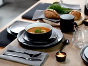 Schwarze Keramikschale mit cremiger Tomatensuppe und Dillgarnitur auf grauen Tellern, daneben schwarzer Becher, Glas, Besteck und Brot auf Holztisch, fotografiert in leicht schräger Perspektive von vorn oben.