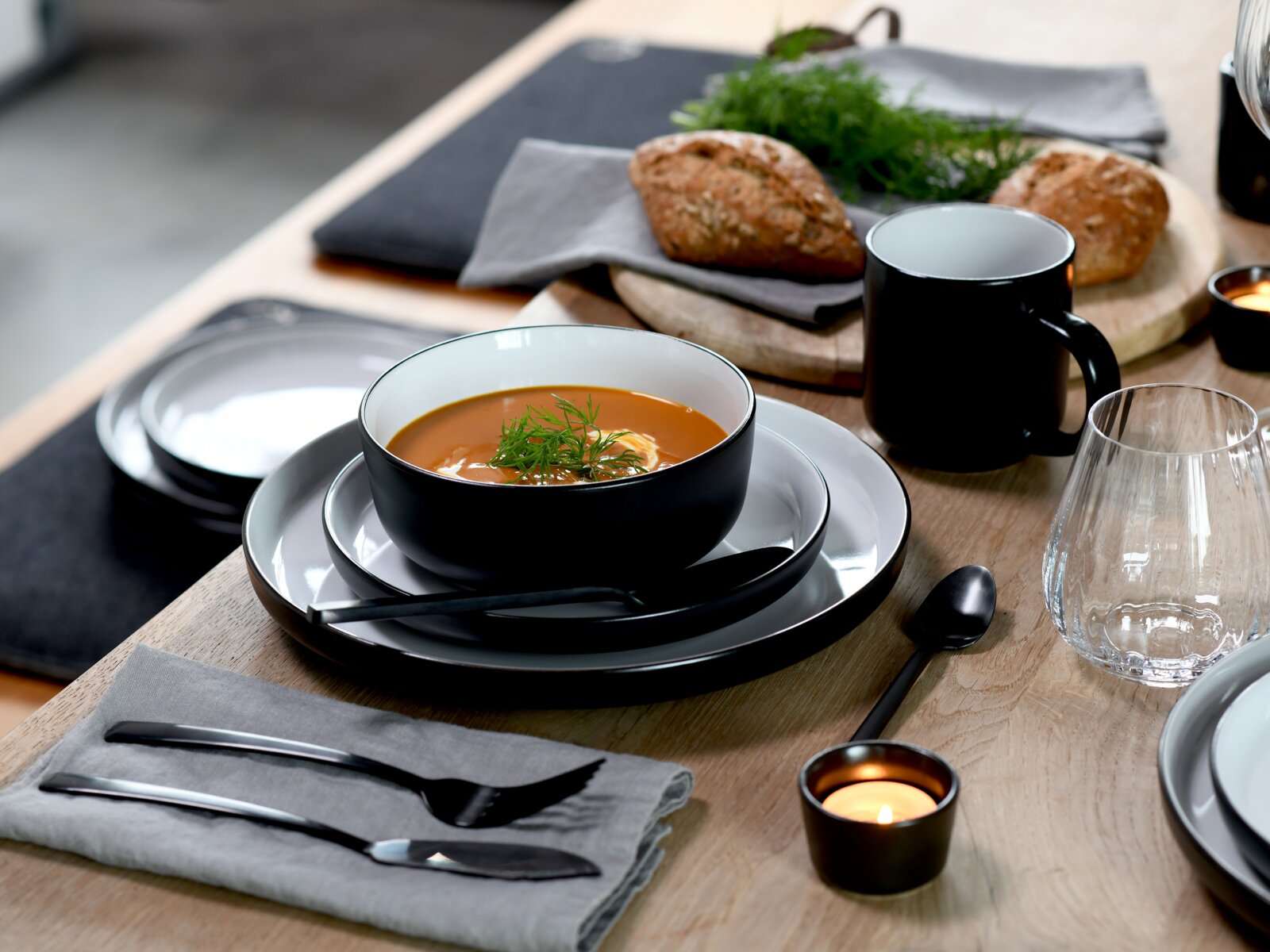 Schwarze Keramikschale mit cremiger Tomatensuppe und Dillgarnitur auf grauen Tellern, daneben schwarzer Becher, Glas, Besteck und Brot auf Holztisch, fotografiert in leicht schräger Perspektive von vorn oben.