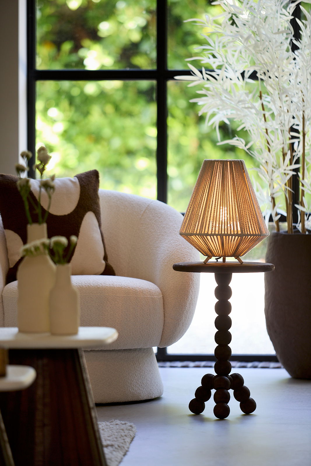 Light&Living Tischleuchte FELIDA Tischleuchte mit geflochtenem Lampenschirm auf einem runden Tisch mit Kugelbeinen, im Hintergrund ein gemütlicher Sessel und ein großes Fenster. Perspektive von vorne.