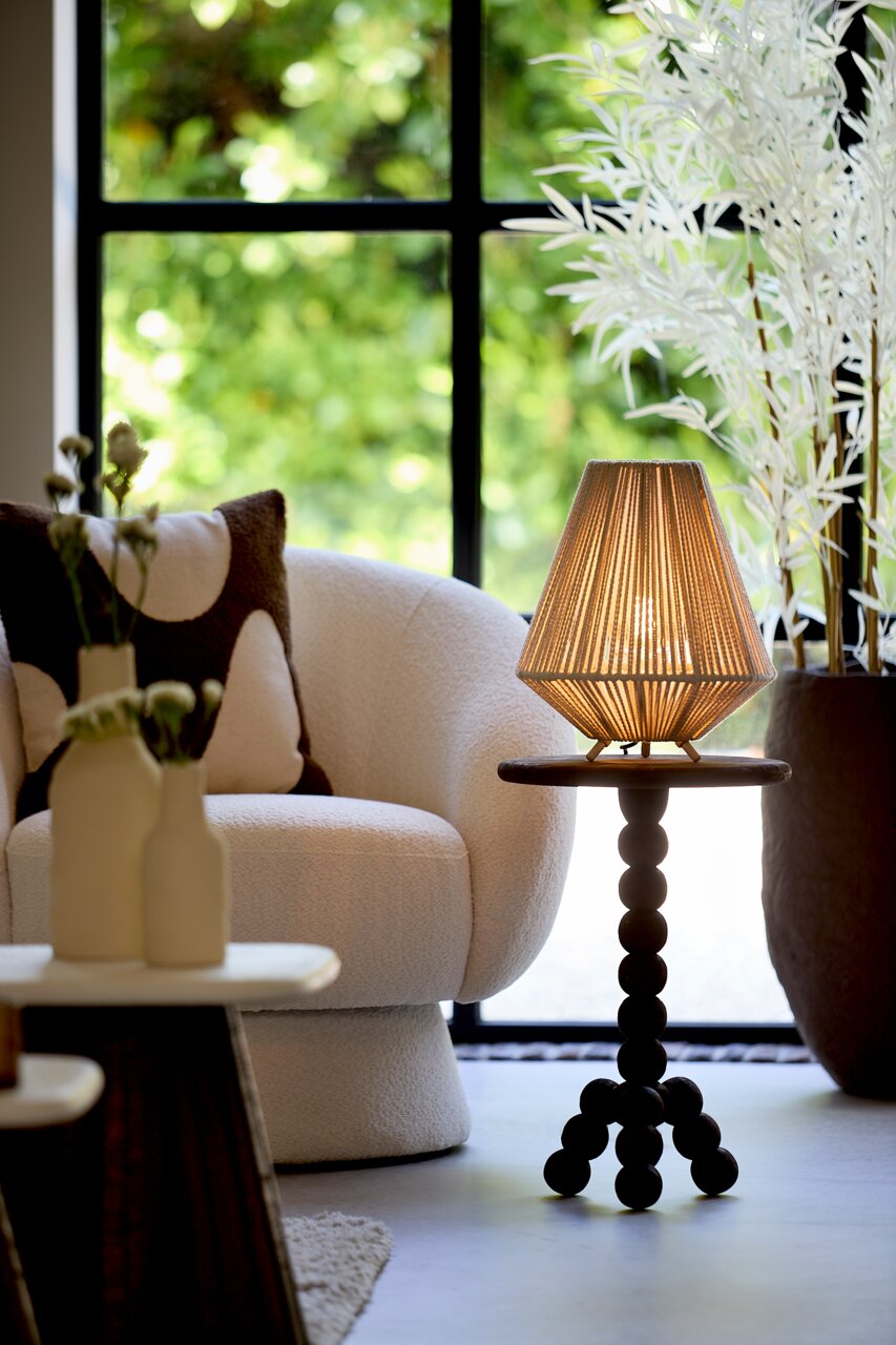 Light&Living Tischleuchte FELIDA Tischleuchte mit geflochtenem Lampenschirm auf einem runden Tisch mit Kugelbeinen, im Hintergrund ein gemütlicher Sessel und ein großes Fenster. Perspektive von vorne.