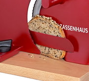 Rote Brotschneidemaschine mit Holzbrett schneidet eine Scheibe Körnerbrot, Nahaufnahme aus seitlicher Perspektive